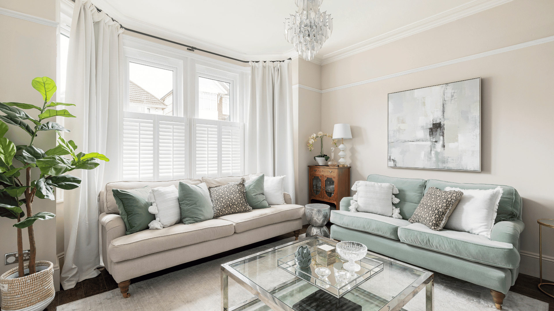 Neutral living room with a bay window dressed in full-length white curtains and shutters, alongside beige and sage green sofas.