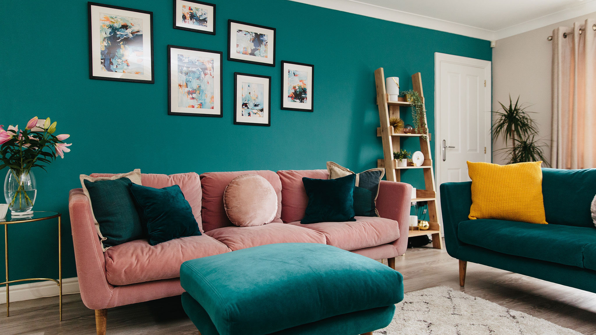 Colourful living room with a teal feature wall, gallery artwork, pink sofa, teal armchair and matching ottoman.