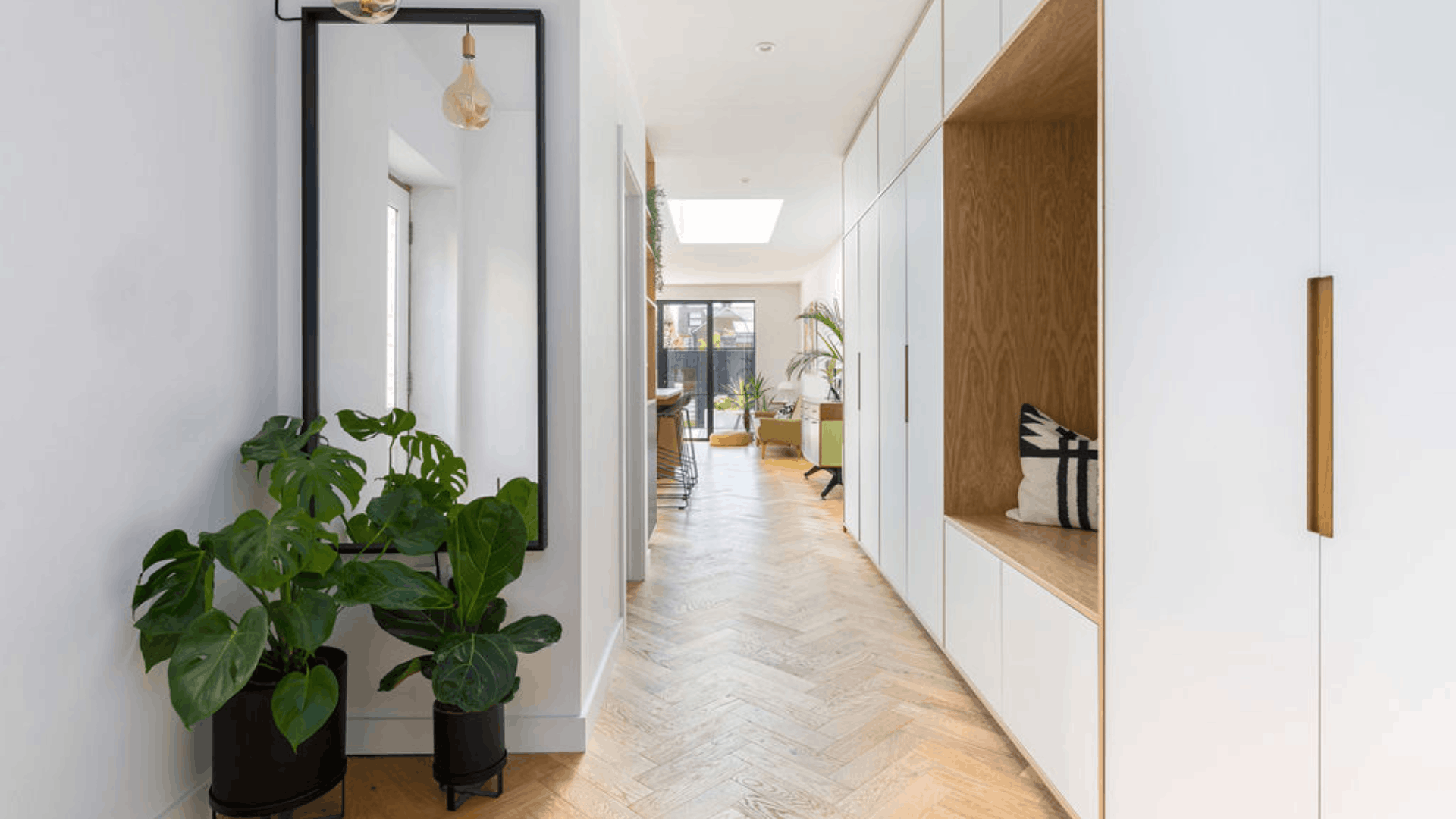 Narrow hallway with a tall mirror, houseplants, white built-in storage and light herringbone flooring.
