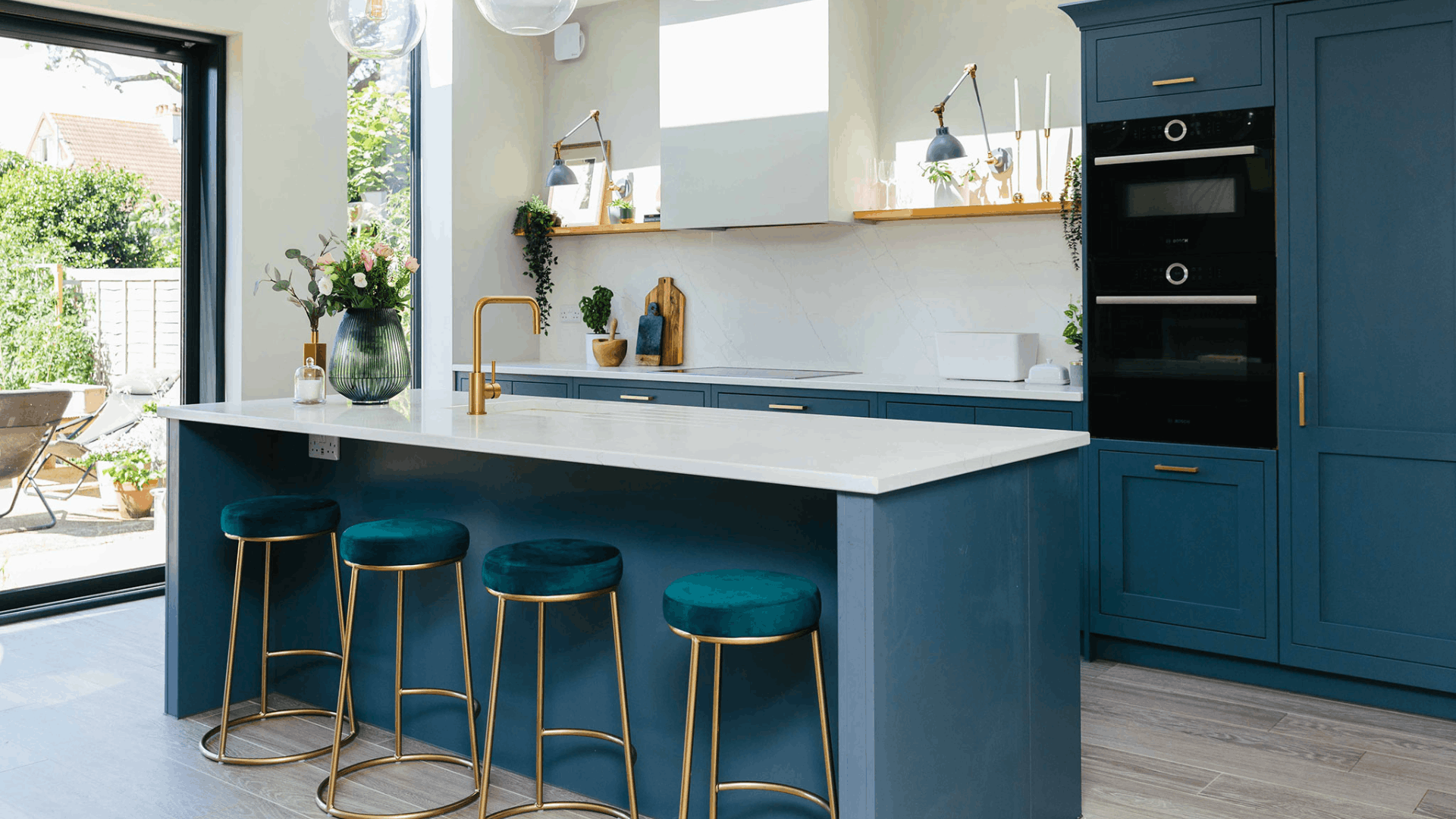 Modern kitchen with painted navy cabinets, a white island worktop, brass handles and teal velvet bar stools.
