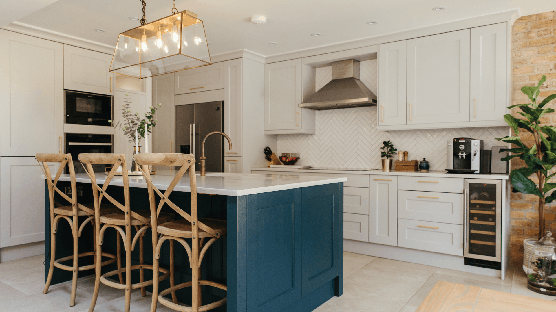 Open kitchen with white shaker cabinets, a navy island, white worktops, wooden bar stools and a brass-framed pendant light.