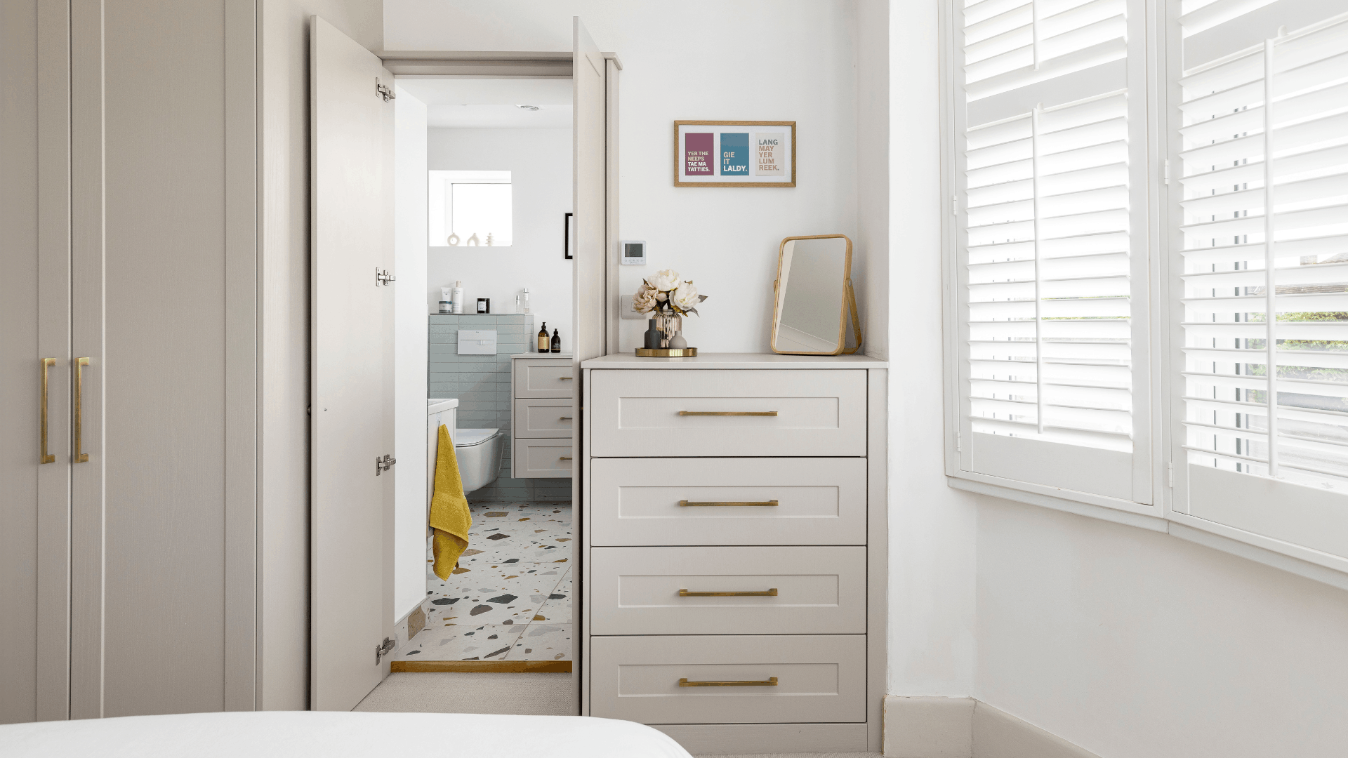 Bright bedroom with built-in wardrobes and drawers, white plantation shutters and an open door leading to an en-suite bathroom.