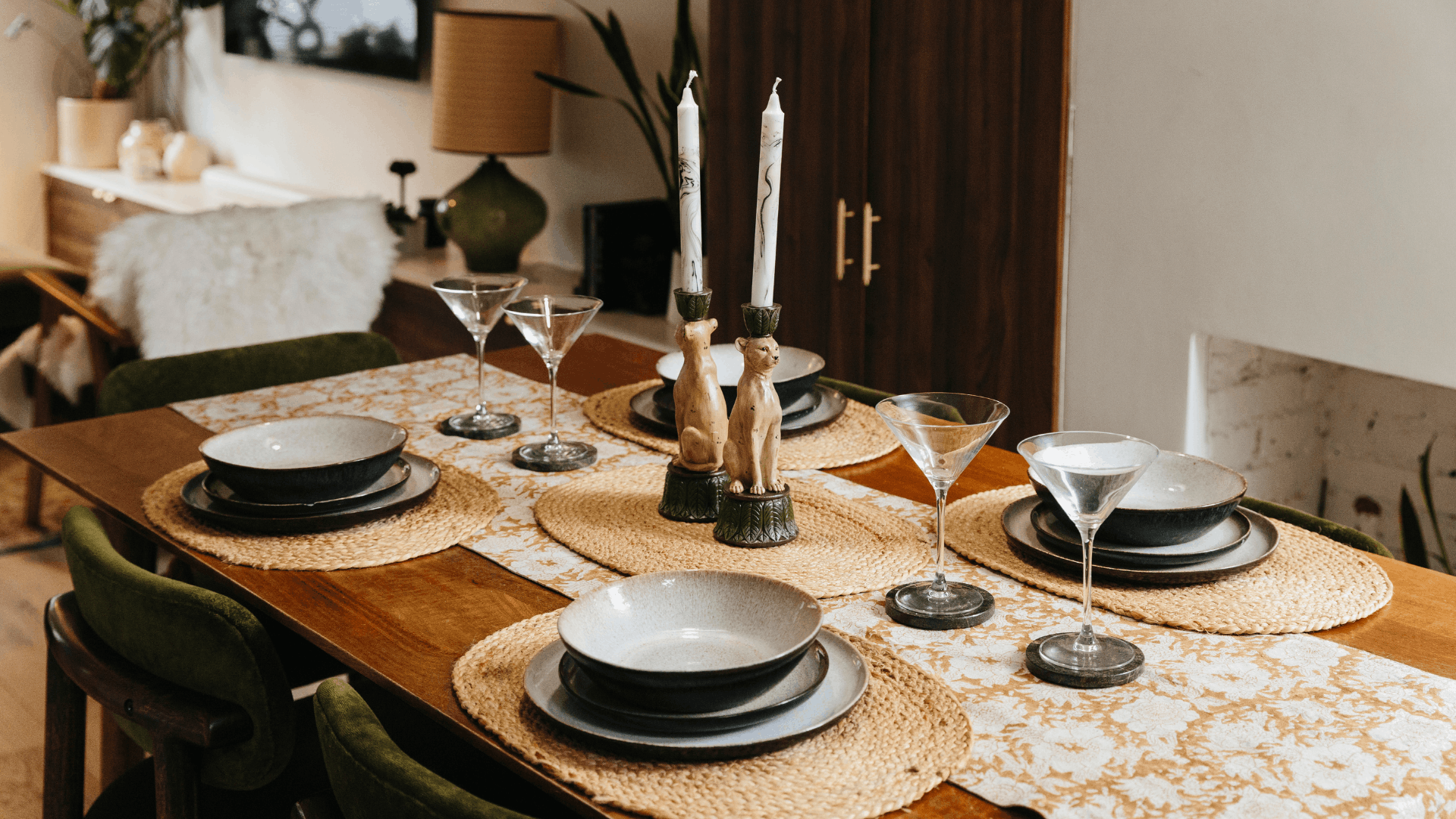 Dining room table with decorative candlesticks, a patterned yellow runner, rattan placemats and olive green dining chairs.