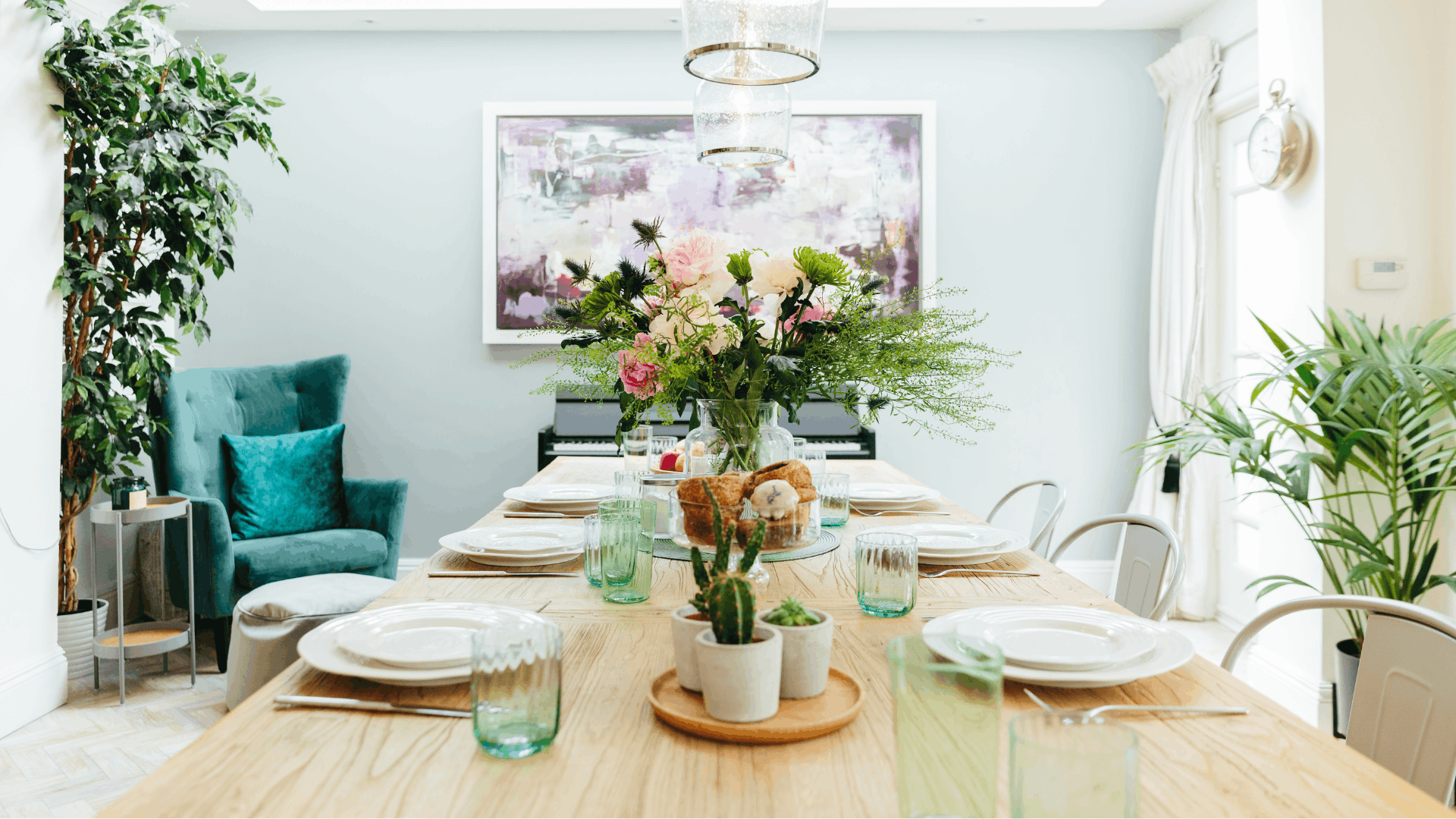 Bright dining room with a wooden table styled with white tableware, green glassware, flowers and small potted plants.