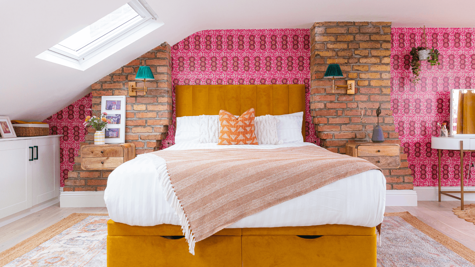 Colourful loft bedroom with slanted walls, exposed brick, pink patterned wallpaper and a mustard upholstered storage bed beneath a skylight.