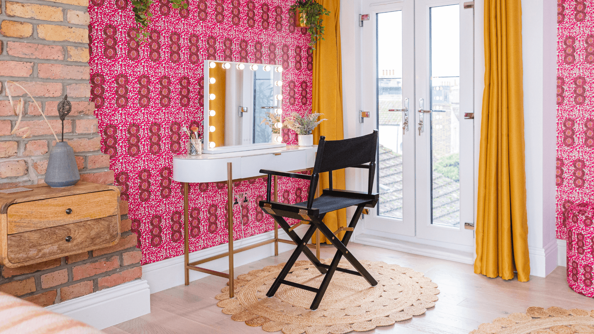 Bedroom dressing area with a light-up vanity mirror, slim dressing table, black chair, mustard curtains and bold pink wallpaper.
